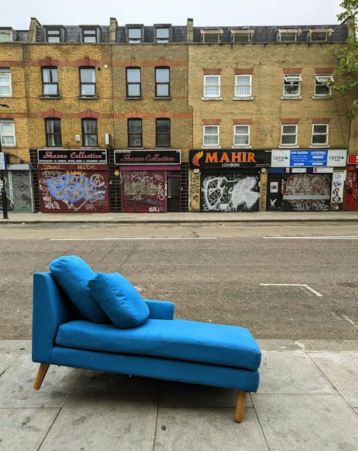 A bright blue modern chaise lounge with a matching cushion and wooden tapered legs is positioned on a sidewalk in front of a brick building. The sofa features a smooth, fabric-covered surface with visible seams and slight tufting on the cushion, offering a contemporary appearance. Behind it, there is a street with an asphalt road, and across the street, a row of storefronts with closed shutters and graffiti, including various tags and murals, is visible. The building above the shops has a brick facade with several windows, some with white frames and curtains, indicating residential or commercial units. The overall scene was likely captured during daylight hours, with natural light illuminating the sidewalk and the furniture. The setting subtly connects to the context of private waste or furniture removal, as the sofa appears to be discarded or awaiting collection, fitting the theme of rubbish clearance or alternative waste disposal methods associated with independent collection services.