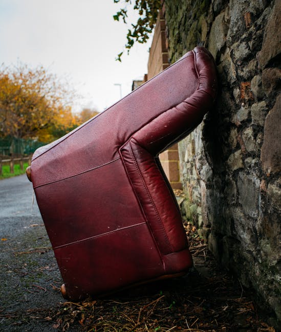 A red leather sofa with a smooth, shiny finish is seen overturned on the ground, leaning against a weathered stone wall in a residential area. The sofa's armrest and seat cushions are visibly creased, and the upholstery shows slight signs of wear, with minor scuffs and scratches. It is positioned on a narrow pavement or alleyway with wet patches, and small twigs and leaves are scattered around its base. In the background, a brick building and a leafless tree are partially visible beneath an overcast sky, indicating an outdoor environment during late autumn. The scene suggests a private collection or on-site clearance of bulky furniture, possibly managed by a waste removal service like rubbishclearancestjohnswood.com, fitting within the broader context of alternative waste handling outside of local authority collection. Natural diffused lighting emphasizes the textures of the sofa’s leather and the roughness of the stone wall, underlining the scene's neutral observational tone and practical focus on waste disposal or furniture removal.