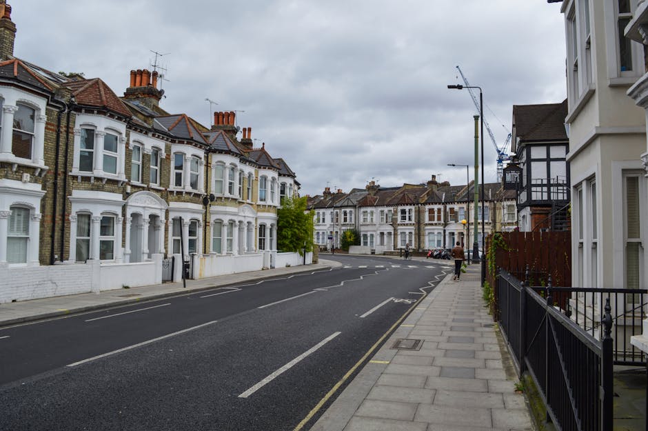 A quiet residential street in St Johns Wood with Victorian-era terraced houses characterized by decorative bay windows, brick facades, and white-painted details. The pavement on the right is lined with a black metal railing and a neatly maintained garden area. Several houses feature chimneys and tiled roofs, with some displaying small front gardens enclosed by low walls. The street is paved with asphalt and marked with white lines indicating parking restrictions; a row of parked cars is visible in the distance. Overhead, a cloudy sky creates diffused natural lighting, and a tall street lamp is mounted along the pavement. This scene exemplifies typical urban architecture, with clear visual cues for independent waste disposal and rubbish collection, as often required by private rubbish removal services in the area. Occasionally, a person can be seen walking along the sidewalk, emphasizing the calm, residential atmosphere of the neighbourhood as a suitable context for professional rubbish removal or on-site clearance solutions.