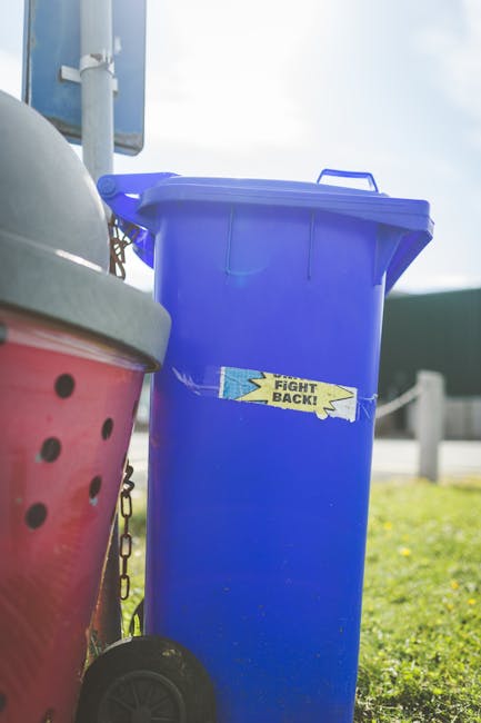 A tall, blue wheeled rubbish bin with a closed lid, positioned outdoors on a grassy area next to other waste containers, including a partially visible pink bin with a perforated design in the foreground. The blue bin, likely made of durable plastic with a smooth surface, stands upright on a pavement or driveway, with sunlight casting a bright, natural light across the scene. A small, torn yellow and blue adhesive label with the words 'FIGHT BACK!' is affixed to the front of the blue bin. In the background, there is a metal pole supporting a sign and a portion of a road or pathway, with a guardrail visible further away. This outdoor setting, characterized by clean, well-maintained waste containers and natural surroundings, subtly reflects the context of independent or private rubbish collection, often associated with alternative waste handling services provided by companies like rubbishclearancestjohnswood.com serving the local area near Lords Cricket Ground.