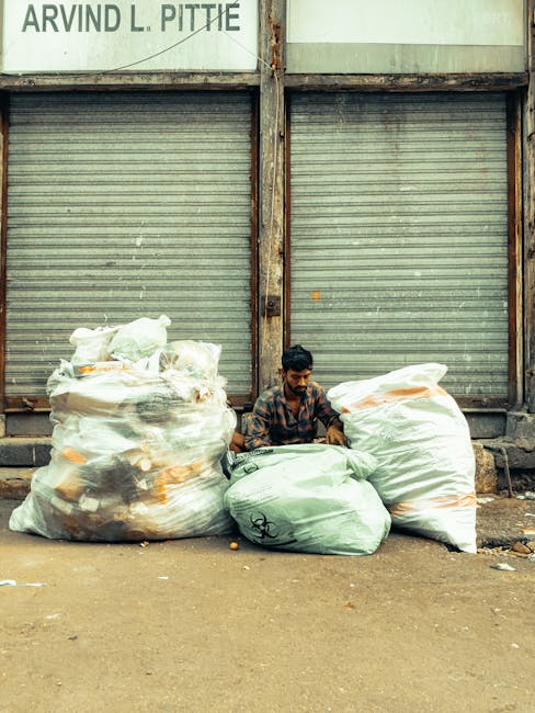 A man sitting on the ground between three large, transparent plastic bags filled with mixed waste and other rubbish, positioned in front of a closed metal shutter with visible rust and dirt marks. The rubbish bags contain various materials, including plastic, paper, and cloth. The setting appears to be an outdoor urban area, possibly a back alley or side street, with a concrete or asphalt surface beneath the man and rubbish bags. The background structure has a weathered appearance, indicating it may be an industrial or commercial building. Natural light illuminates the scene evenly, emphasizing the textures of the waste materials and the gritty environment. This visual context relates to private waste collection or rubbish clearance services provided by companies specializing in on-site clearance or alternative waste handling solutions in urban settings, highlighting the need for proper rubbish removal in such environments.