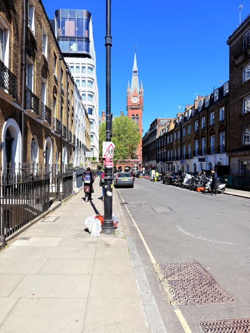 A daytime street scene in an urban area featuring a clear blue sky and a prominent red-brick clock tower with a tall spire in the background. The foreground shows a sidewalk with railing, and a black lamppost positioned near the center. On the sidewalk, there is a white plastic bag filled with rubbish placed next to a traffic sign attached to the lamppost. The street is lined with multi-storey terraced buildings on both sides, exhibiting brick facades, arched windows, and chimneys, with some buildings displaying white window frames and balconies. Parked vehicles, including cars and motorcycles, are visible along the curb. Pedestrians are walking along the sidewalk, and the scene captures an atmosphere of urban everyday activity, with visible rubbish and waste collection bins indicating ongoing waste management activities. The overall scene reflects an area where independent waste clearance services like those from rubbishclearancestjohnswood.com could operate to assist with rubbish removal and alternative waste disposal outside of local authority services.