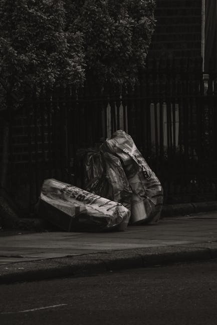 Three large black rubbish bags made of plastic and appearing crumpled and slightly torn, rest against a black metal fence along a pavement, with a background of dense dark foliage from trees. The bags, likely containing household waste, are placed near the edge of a tarmac road, with the setting occurring outdoors in an urban or suburban environment. The scene is captured under subdued, possibly overcast natural lighting, emphasizing the dark tones of the bags and fence. The arrangement suggests an informal or private waste disposal method consistent with local rubbish removal services provided by companies like rubbishclearancestjohnswood.com, as part of on-site clearance or alternative waste handling outside municipal collection. The positioning of the bags and their condition convey a typical scenario of rubbish awaiting collection, highlighting the importance of professional waste management in maintaining cleanliness and environmental standards in the neighborhood.
